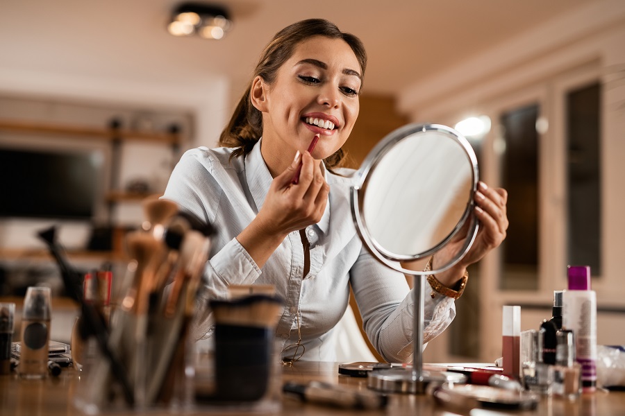 Young smiling woman applying lip liner while looking herself in a mirror.